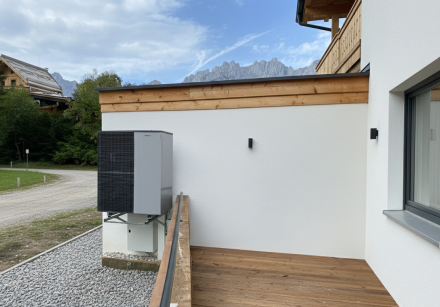 Eine hochmoderne Wärmepumpe ist an der Außenwand eines modernen Hauses installiert, das über eine Holzterrasse und eine beeindruckende Bergkulisse verfügt. Die effiziente Heizlösung ist ideal für nachhaltiges Wohnen und Energieeffizienz. waermepumpe