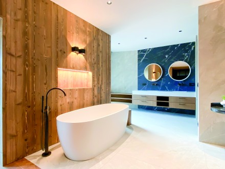 Modernes Luxus-Badezimmer mit freistehender Badewanne, stilvoller Holzwand, elegantem Waschtisch mit Marmorrückwand und runden Spiegeln – Design trifft auf Wohlfühl-Atmosphäre. bad_und_sanitaer